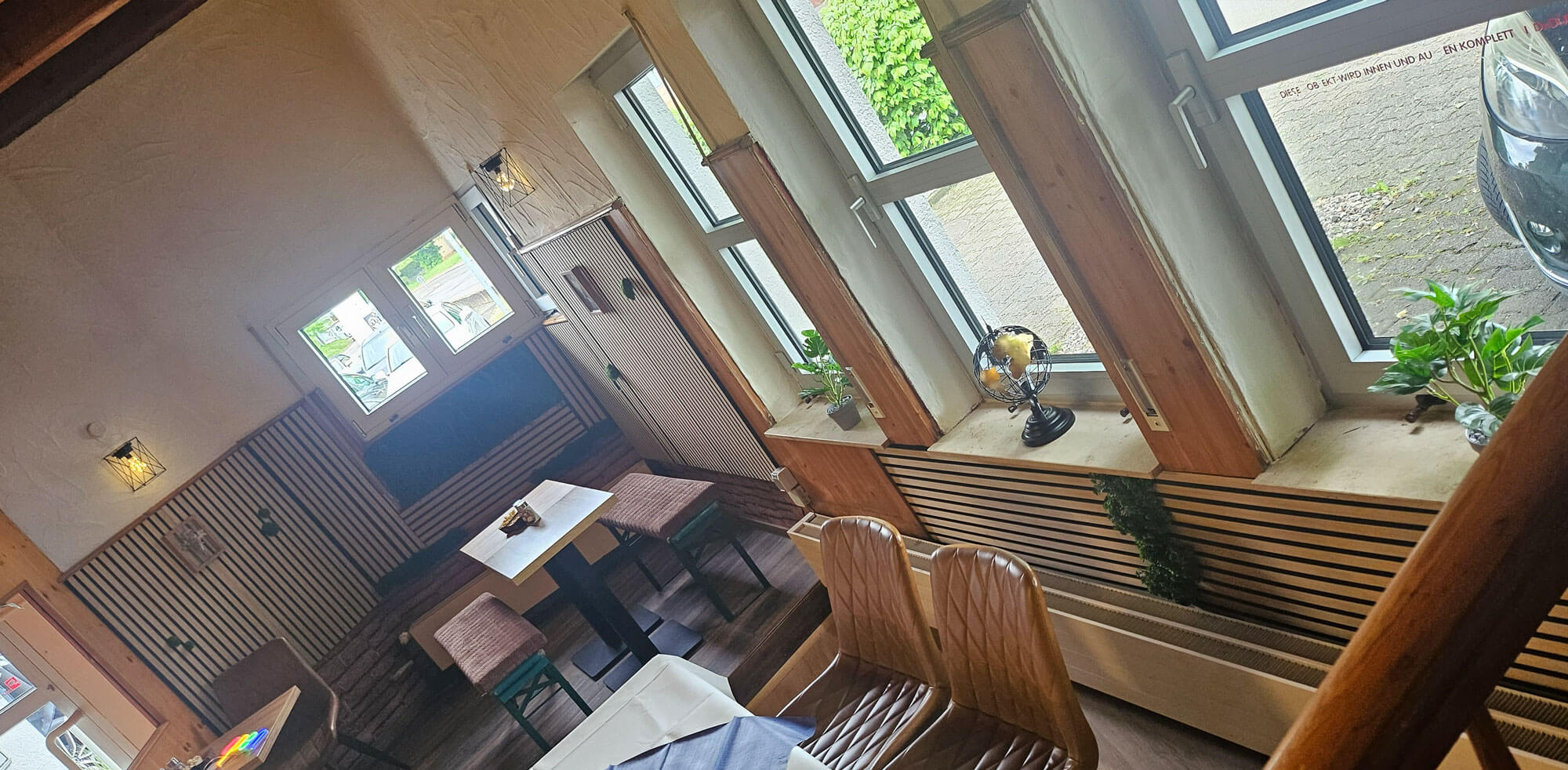 Leerer Gastraum mit ruhigen Holzbänken in Braun und großen Fenstern, warmes Licht und Pflanzen am Fensterbrett